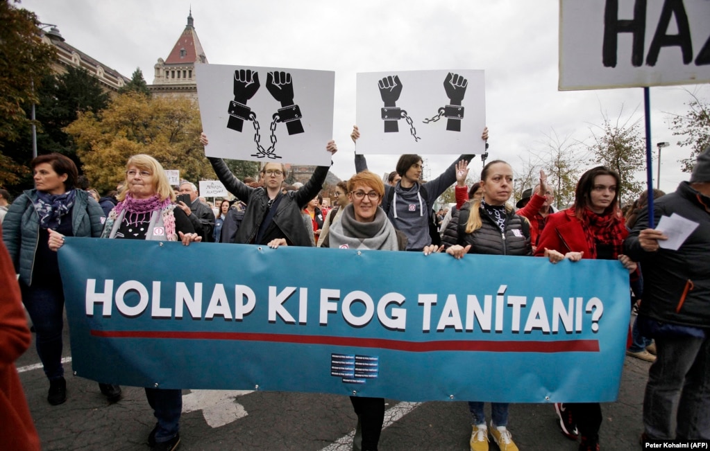 Gratë mbajnë një pankartë ku pyesin: &quot;Kush do të jap mësim nesër?&quot; Që nga fillimi i vitit shkollor 2022, mësuesit dhe studentët kanë mbajtur demonstrata në Budapest dhe gjetkë për të treguar mbështetje për mësuesit e shkarkuar për shkak të bashkimit me protestat e mëparshme. &nbsp;