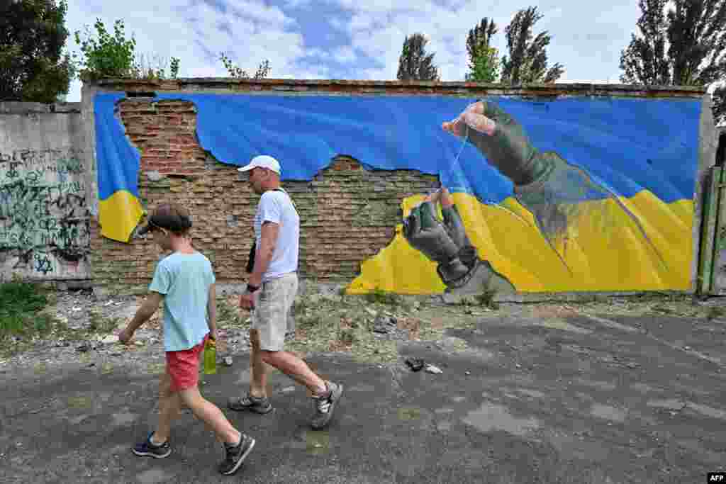 Vendasit kalojnë pranë një murali që tregon duart e një ushtari ukrainas duke riparuar flamurin e grisur të Ukrainës në Kiev. Fotografuar në prill. &nbsp;