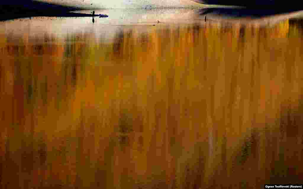 A fisherman stands on the bank of North Macedonia&#39;s Globocica Lake with autumn foliage in the background.