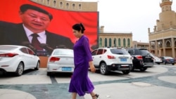 Женщина-уйгурка на улице в Кашгаре на фоне гигантского монитора с трансляцией выступления главы Китая Си Цзиньпина. 2018 года
