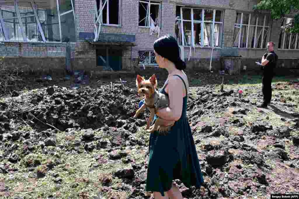 A woman holding a dog stands by a huge crater from a rocket strike close to a destroyed school building in Kharkiv on July 4.