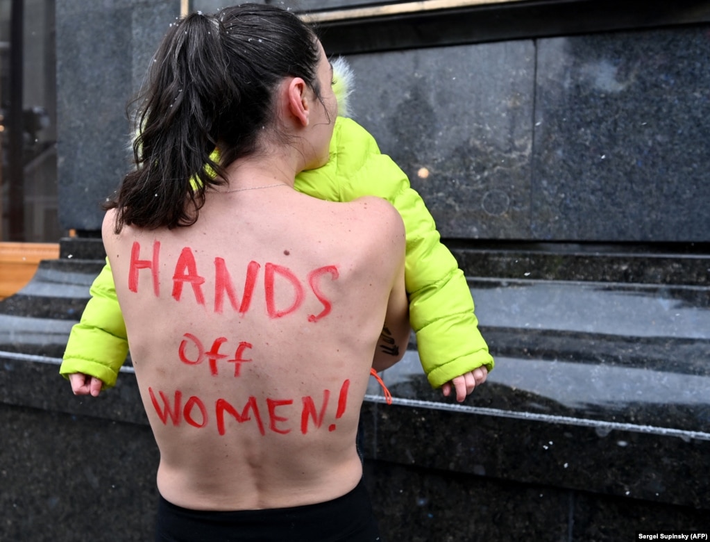 Një aktiviste e grupit 'FEMEN' duke protestuar me vajzën e saj të mitur jashtë zyrës së presidentit të Ukrainës, më 27 janar. Gruaja, e zhveshur në pjesën e epërme të trupit, po protestonte ndaj një rregulloreje që hyri në fuqi në fund të vitit 2021, e cila u kërkon grave të moshës 18-60 vjeç nga një sërë profesionesh që të regjistrohen në ushtrinë e Ukrainës. Në rast të një pushtimi, gratë mund të mobilizohen për të ndihmuar në një përpjekje lufte.