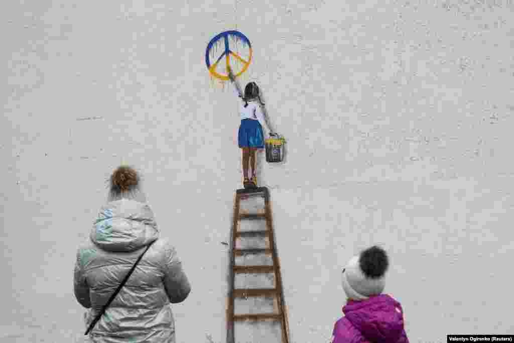 Жінка з дитиною дивляться на зображення дівчинки, яка малює антивоєнний знак. Цей образ створив вуличний художник Tvboy на стіні ліцею в Бучі. Україна, 29 січня 2023 року