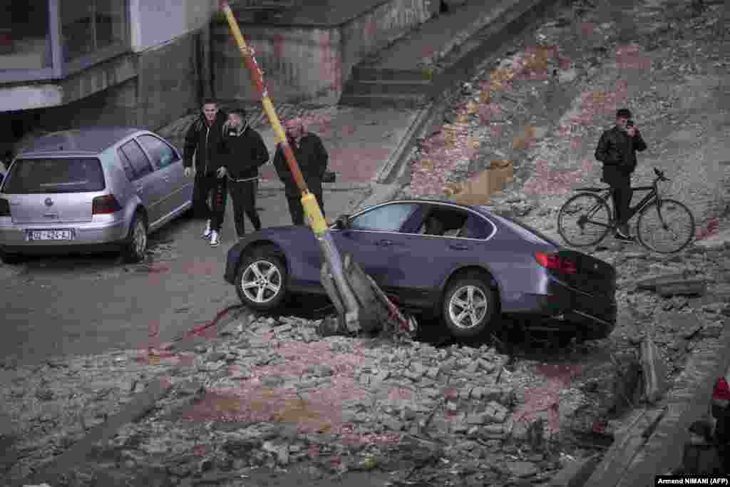 Pedestrians walk by a damaged and displaced car on a street after heavy rain in Skenderaj, Kosovo.