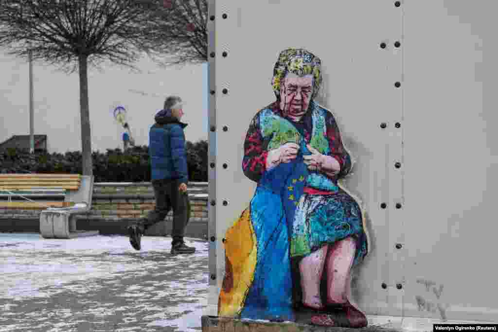 A man walks by a mural in Bucha of an older woman stitching together an EU and Ukrainian flag. The picture is outside a makeshift aid station that provides heating and electricity during blackouts -- so-called &quot;points of invincibility.&quot;&nbsp;&nbsp; &nbsp;