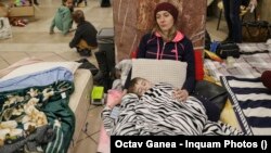 O tânără refugiată din Ucraina se odihnește în sala de așteptare din Gara de Nord din București alături de băiețelul ei, în timp ce alți copii refugiați se joacă