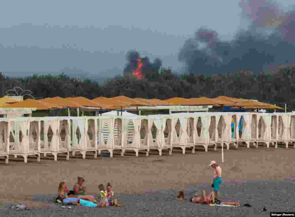 People relax on a beach as smoke and flames rise after explosions at a Russian military air base, in Novofedorivka, in Ukraine&#39;s occupied Crimea region, on August 9.
