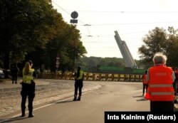 Rendőrök és a média figyeli, ahogy ledöntenek egy nyolcvan méter magas obeliszket – egy második világháborús szovjet győzelmi emlékművet – Rigában 2022. augusztus 25-én