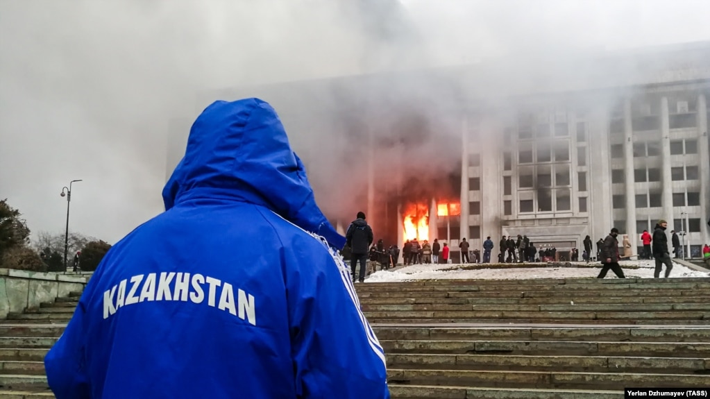 Человек в куртке с надписью: «Казахстан» — напротив горящего здания акимата Алматы 5 января 2022 года