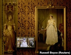 Mbretëresha Elizabeth II në ekranin e televizionit dhe portreti i saj i vendosur në mur. Ndërkaq, në pjesën e majtë të fotografisë shihet statuja e Mbretëreshës Elizabeth I, një prej monarkeve më të shquara britanike.