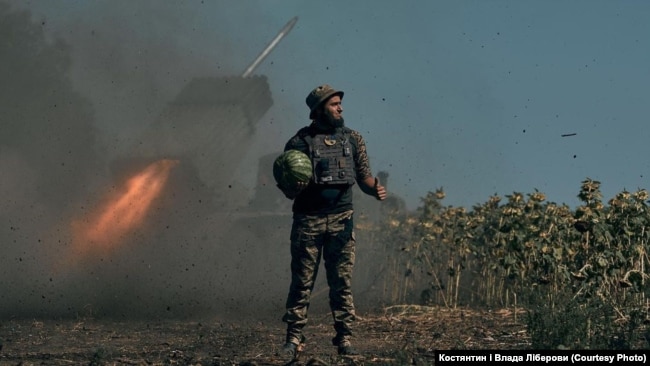 Një ushtar ukrainas mban një shalqi në dorë, përballë një raketehedhëse që gjuan në pozicionet e armikut. Fotografi nga Vlada dhe Kostyantyn Liberov.