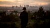 Американец с бутылкой вина на холме в центре Лос-Анджелеса наблюдает закат солнца в дни эпидемии. 23 марта 2020 года