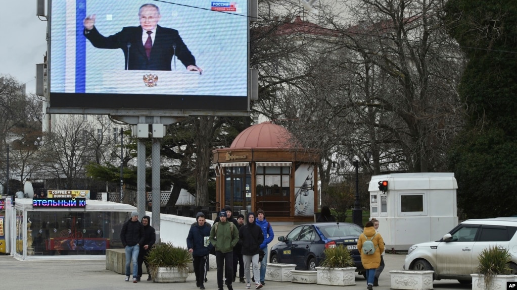 Az elcsatolt Krímben lévő Szevasztopolban emberek sétálnak a februári évértékelő beszédét tartó Vlagyimir Putyin orosz elnököt mutató hatalmas utcai monitor előtt