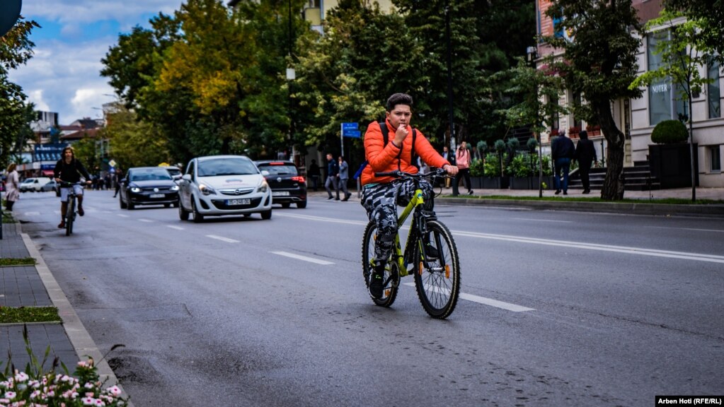 Një djalë i ri duke ngarë biçikletën në rrugët e Prishtinës, ndërsa prapa tij shihen vetura duke qarkulluar.&nbsp;
