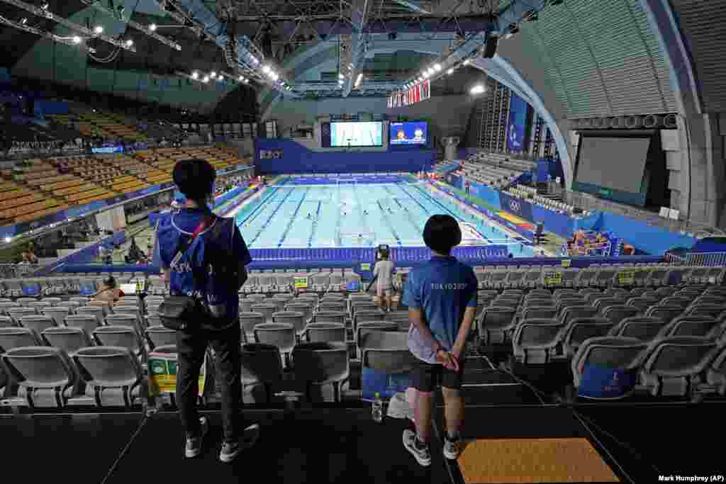 Два волонтера наблюдают за матчем предварительного раунда мужского водного поло из-за пустых трибун в Центре водного поло Тацуми на летних Олимпийских играх 2020 года, в воскресенье, 25 июля 2021 года, в Токио, Япония