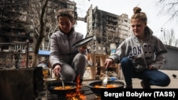 În imagine, femei ucrainence își gătesc în stradă, în aprilie 2022, în Mariupol, lângă un bloc distrus de bombardamente. A fost unul din cele mai afectate orașe de războiul declanșat de Rusia în Ucraina.