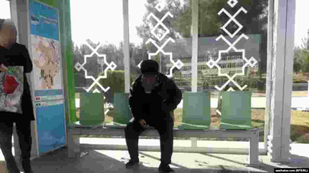 Aşgabadyň başga bir awtobus duralgasynda oturgyçda oturan adam we dik aýak üstünde duran zenan.