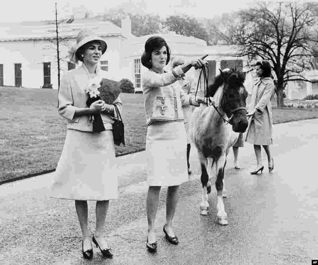 На этой фотографии, сделанной в апреле 1962 года, первая леди США Жаклин Кеннеди проводит экскурсию по территории Белого дома для императрицы Фарах Пехлеви. Кеннеди ведет пони своей дочери Кэролайн, Макарони, которая нюхает императрицу, привлеченная нарциссами в ее руках. На заднем плане &mdash; пресс-секретарь первой леди Памела Турнюр