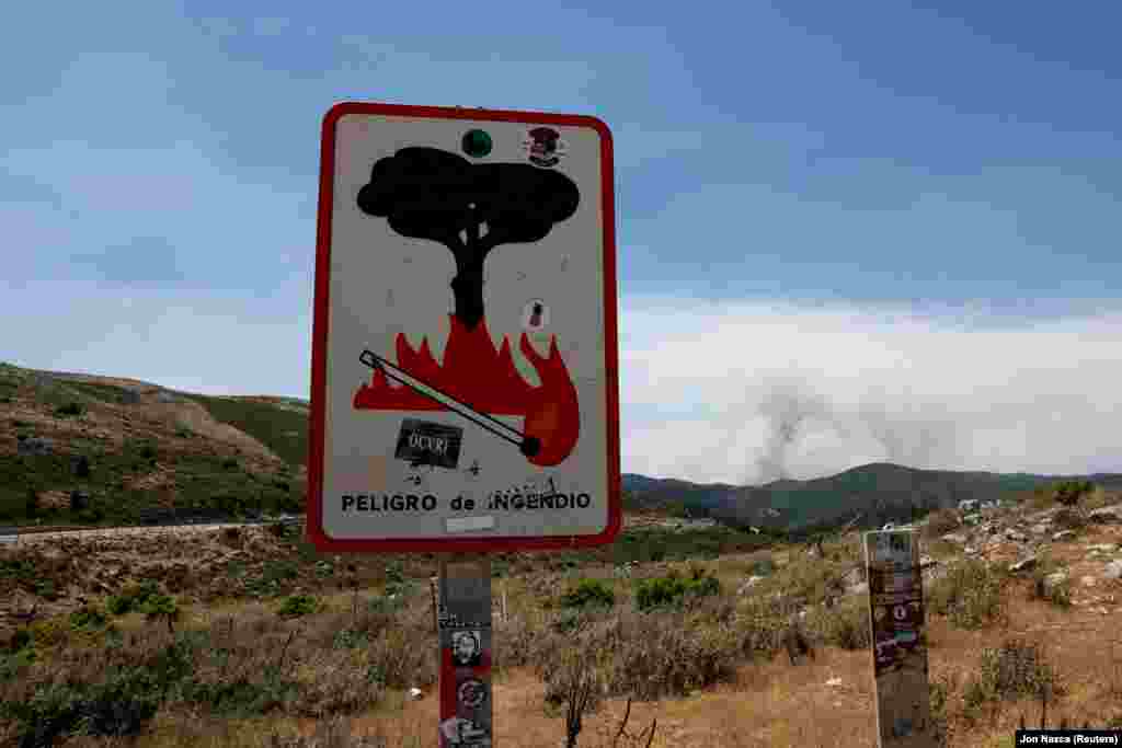 Tűzgyújtási tilalomra figyelmeztető tábla Igualeja közelében. A háttérben erdőtűz pusztít. A tűzvészben eddig három tűzoltó szenvedett égési sérüléseket, egyikük testének 25 százaléka megégett