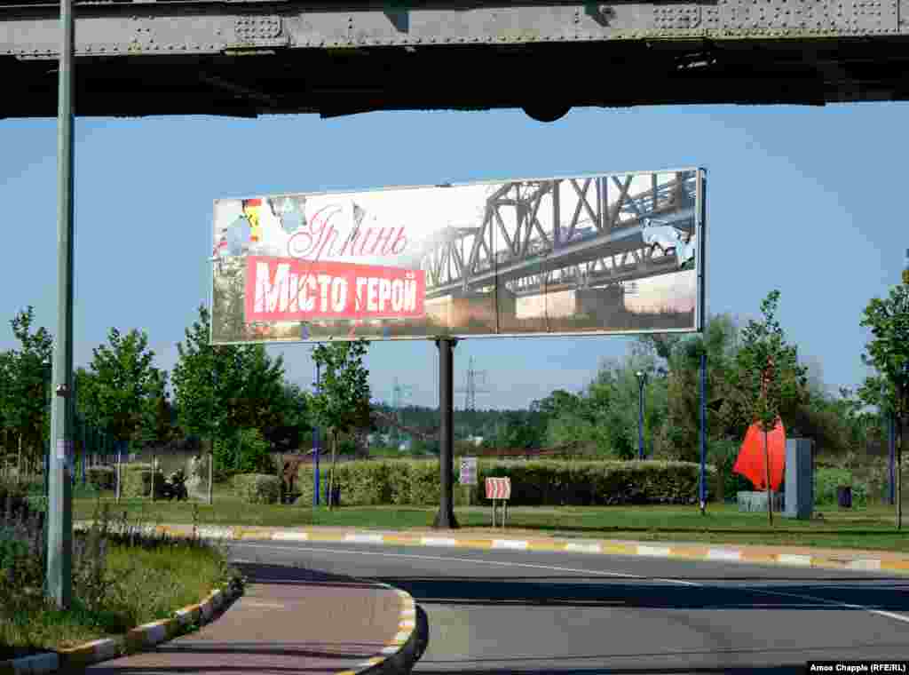 &quot;Irpin, Hero Town&quot; -- next to the Irpin River. One of the two railway bridges on the billboard is visible at the top of this image. Both bridges were destroyed during the opening days of the Russian invasion, but one has already been repaired. A soldier prevented pictures of the bridge being taken when RFE/RL visited.