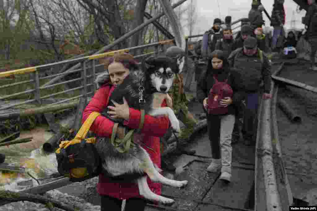 O femeie ține în brațe un câine în timp ce traversează râul Irpin pe o potecă improvizată de sub un pod.&nbsp;Fotografie din 5 martie 2022.