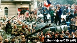 Пражани перекинули автівку, перегородивши дорогу танкам сил Варшавського договору. Радянські солдати з понурими обличчями сидять зверху на броні в оточенні прихильників Празької весни. Чехословаччина, 21 серпня 1968 року
