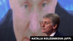 Dmitrij Peskov sjedi ispred ekrana na kojem se prikazan ruski predsjednik Vladimir Putin.