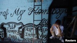 Član ulične bande "Calle 18" pokriva lice dok čuči pored zida na kojem su grafiti ispisani u sjećanje na poginule članove ove bande u zatvoru San Pedro Sula, Honduras, 2013. 