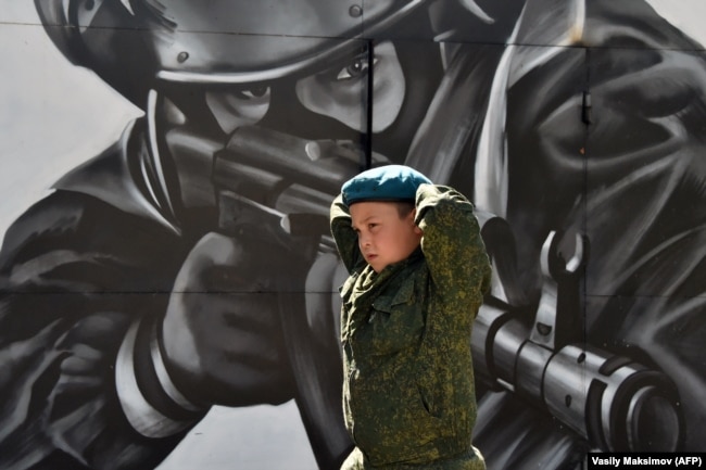 Një djalë i vogël me rroba kamuflazhi duke kaluar pranë një murali ku shfaqet një pjesëtarë i forcave speciale gjatë një gare të llojit ushtarak, të organizuar nga forcat e sigurisë së Gardës Kombëtare për kadetët dhe të rinjtë, në një qendër trajnimi në periferi të Moskës.