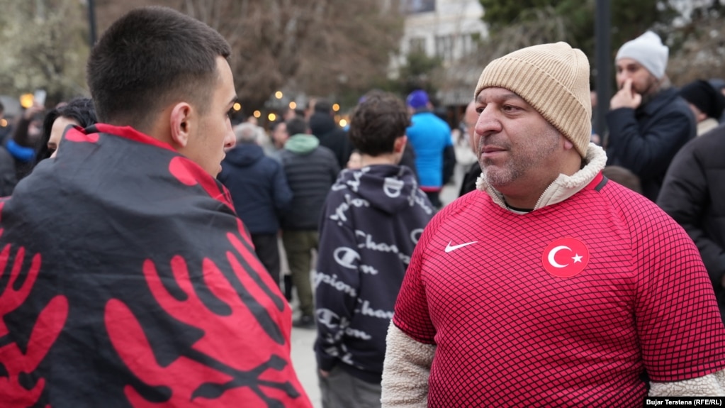Një tifoz i mbështjellë me flamur kombëtar shqiptar dhe një tifozë me fanellë të kombëtares së Turqisë shikojnë njëri-tjetrin në sheshin e Prishtinës, para ndeshjes vendimtare mes Kosovës dhe Turqisë për një vend në Kupën e Botës 2026, të martën në Stadiumin "Fadil Vokrri".