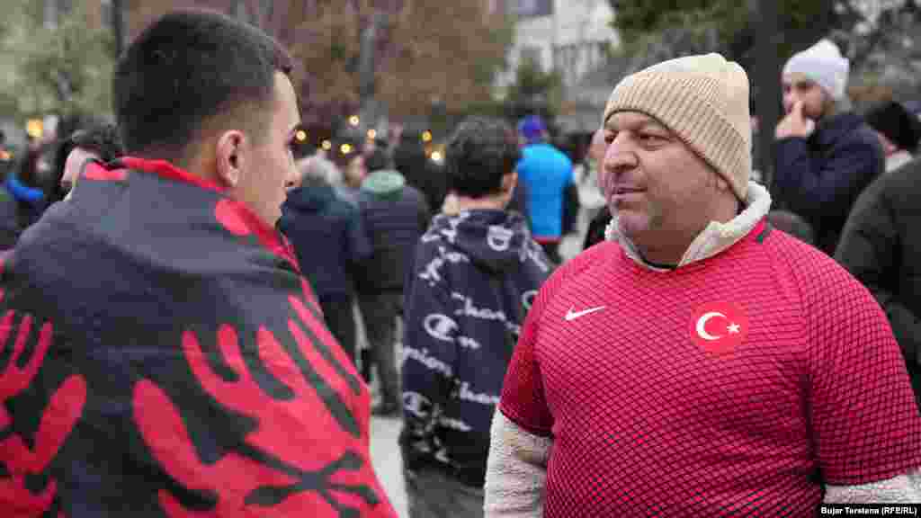 Një tifoz i mbështjellë me flamur kombëtar shqiptar dhe një tifozë me fanellë të kombëtares së Turqisë shikojnë njëri-tjetrin në sheshin e Prishtinës, para ndeshjes vendimtare mes Kosovës dhe Turqisë për një vend në Kupën e Botës 2026, të martën në Stadiumin "Fadil Vokrri".