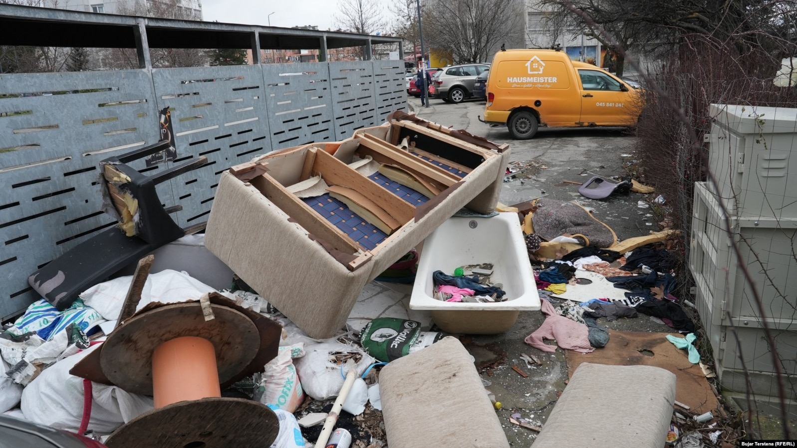 Një vaskë dhe pjesë të një shtrati shihen prapa kontejnerëve në një zonë të Prishtinë.