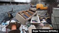 Një vaskë dhe pjesë të një shtrati shihen prapa kontejnerëve në një zonë të Prishtinë.