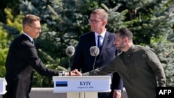 ფინეთის პრეზიდენტი ალექსანდრ სტუბი ხელს ართმევს უკრაინის პრეზიდენტ ვოლოდიმირ ზელენსკის უკრაინის სახელმწიფო დროშის დღისადმი მიძღვნილ ცერემონიაზე. შუაში  ესტონეთის პრემიერმინისტრი კრისტენ მიხალი. კიევი, 2026 წლის 23 აგვისტო
