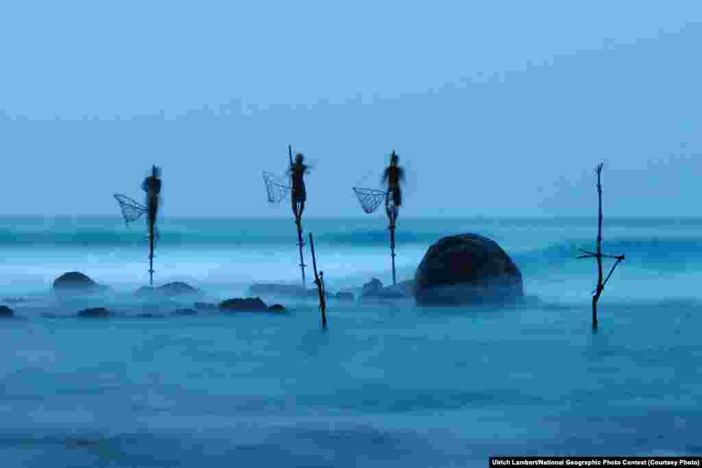 HONORABLE MENTION: Stilt Fishing (Midigama, Sri Lanka) -- Stilt fishing is a typical fishing technique only seen in Sri Lanka. The fishermen sit on a cross bar called a petta tied to a vertical pole planted into the coral reef. This long exposure shot shows how unstable their position is. (Caption by photographer Ulrich Lambert)