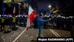 Чоловік із державним прапором Польщі позує перед поліцейським кордоном, тоді як протестувальники збираються біля будівлі Конституційного суду Польщі у Варшаві — після того, як 31 жовтня 2026 року поліція перекрила доступ до цієї будівлі