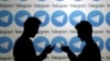 Two men pose with smartphones in front of a screen showing the Telegram logos - generic