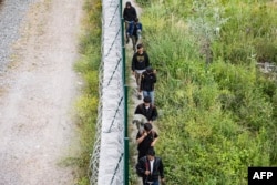 Migranți merg de-a lungul unui gard de sârmă ghimpată în timp ce părăsesc o tabără improvizată, în urma unei operațiuni de demontare desfășurate de poliția franceză la Dunkerque, în nordul Franței, în iunie 2025
