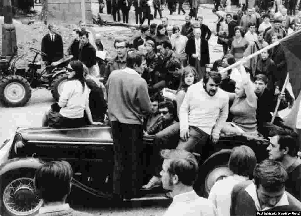 This picture has so many contrasting elements, the clearly overloaded sports car, driven by a Bob Dylan look-alike wearing dark glasses, a flag waving, mini-skirted girl and a tractor in the background.