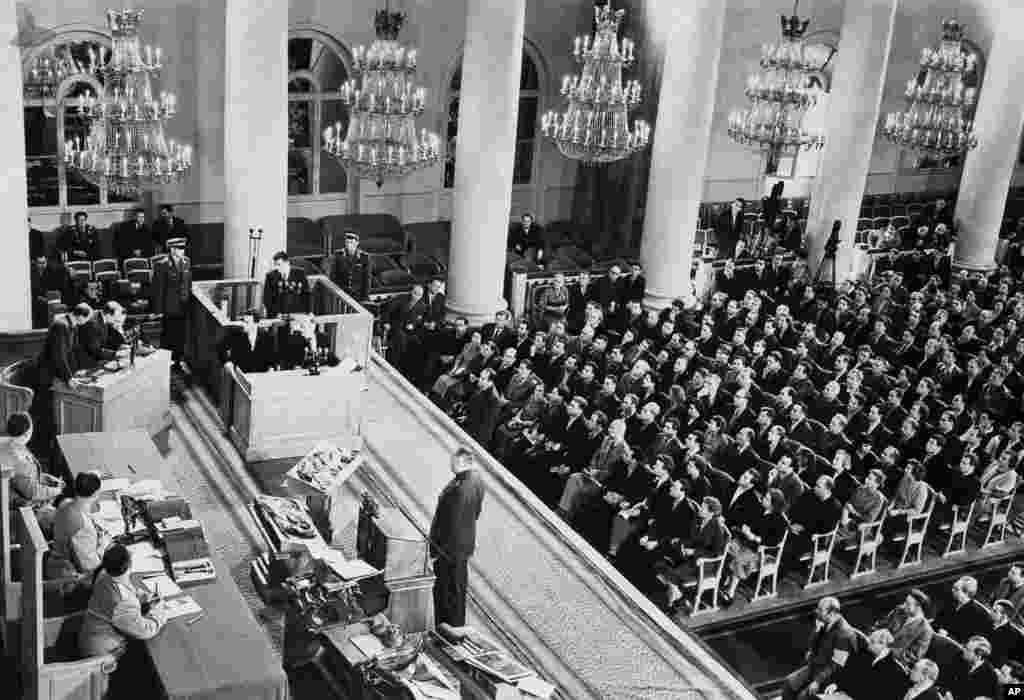 Powers sits in the dock in the Hall of Columns. Immediately in front of Powers is his defense counsel, Mikhail Griniev. In the foreground at left are the judges: three high-ranking Soviet officers. Just in front of their bench is equipment said to have been carried by Powers in his plane.