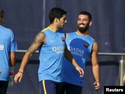 Arda Turan (j) Luis Suárezzel a Barcelona színeiben viccelődik edzés közben a katalán csapat edzőpályáján 2015. július 13-án – de a logó ironikus módon Katarra utal