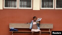 Një vajzë lexon një libër, teksa qëndron e ulur në një stol pranë një shkolle në Mozambik.
