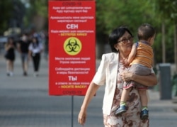 Карантин кезінде көшедегі COVID-19 туралы ақпарат жазылған баннердің қасынан өтіп бара жатқан бала көтерген әйел. Алматы, 12 шілде 2020 жыл.