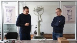 Vladimir Melnic (stânga), vicedirector la Centrul Național Tehnologii Spațiale de la UTM și Alexei Martîniuc, cercetător științific, discutând pe fundalul prototipului unui satelit lansat anterior în spațiu.