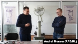 Vladimir Melnic (stânga), vicedirector la Centrul Național Tehnologii Spațiale de la UTM și Alexei Martîniuc, cercetător științific, discutând pe fundalul prototipului unui satelit lansat anterior în spațiu.