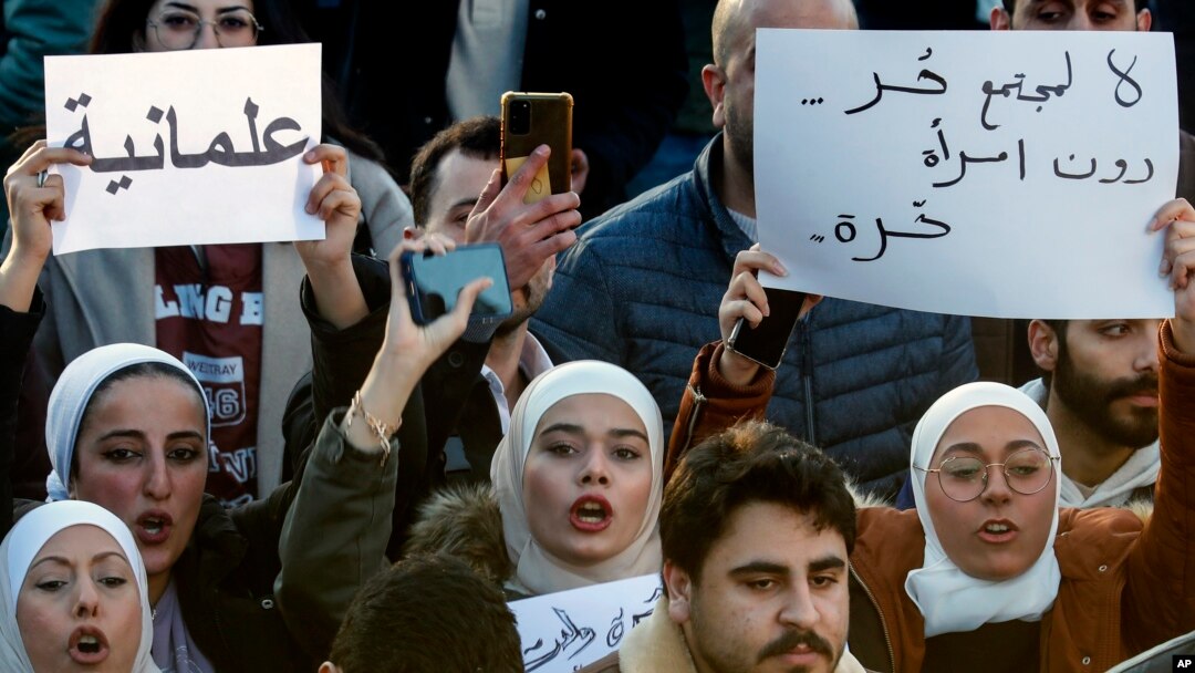 Femei siriene scandând sloganuri și ținând în mâini pancarte în limba arabă pe care scrie: „Nu există societate liberă fără femei libere” (dreapta) și „Secularism” (stânga), la o manifestație de revendicare a unui stat secular, Damasc, Siria, 19 decembrie 2024.