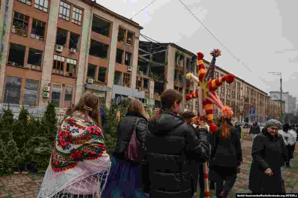 Різдвяна хода проходить повз вуличний ринок, де продають ялинки, на тлі зруйнованої адміністративної будівлі неподалік від станції метро «Лукʼянівська»