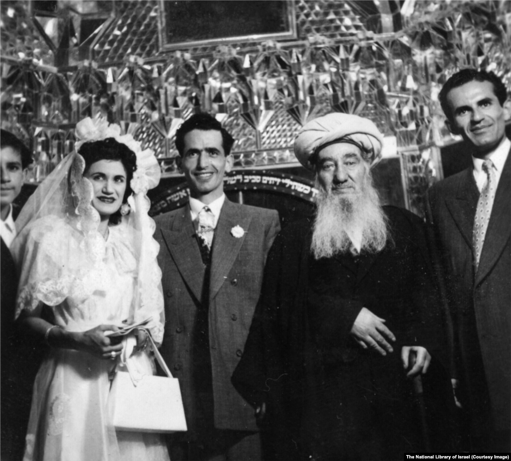 A Jewish wedding at an unspecified location in Iran in the 1950s. The ceremony is being officiated by Rabbi Shlomo Cohen Tzedek (center right), the chief rabbi of the country.Hebrew script behind the group references the bible verse: “Your wife will be like a fruitful vine within your house; your children will be like olive shoots around your table.”