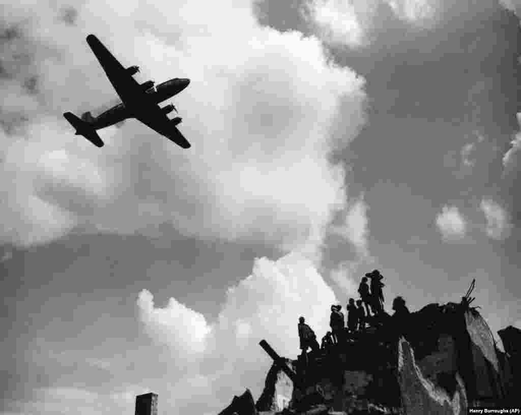 Tineri stau pe o clădire disatrusă de bombe, lîngă Tempelhof, în timp ce un avion american zboară deasupra capetelor lor, după de livrase o încărcătură de cărbune la 9 iulie 1948. Pentru ca orașul să supraviețuiască blocadei sovietice, s-a estimat că berlinezii aveau nevoie zilnic de cel puțin 4500 de tone de alimente și cărbune. Coordonarea eficientă a zborurilor le-a permis aliaților să depășească treptat acest minim.