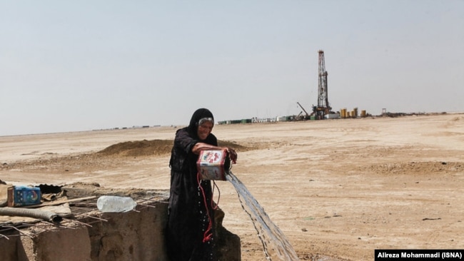 Gheyzaniyeh -- A local woman takes water from a well while in the background an oil well is visible.